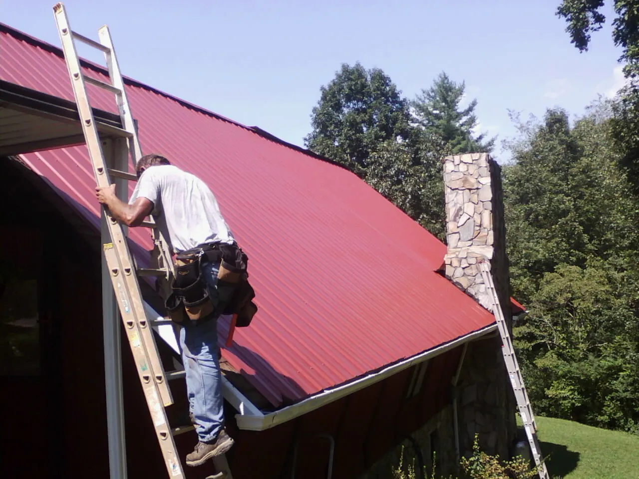 Roofer on a ladder beside a red metal roof and stone chimney — Soffit & Fascia Repair by Roofing Brothers in Hilliard